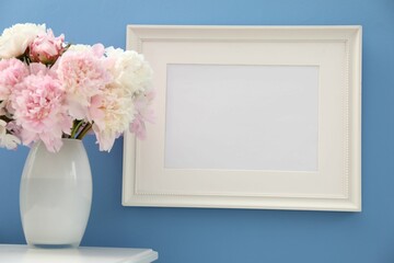 Beautiful peonies in vase and picture frame on white table near light blue wall