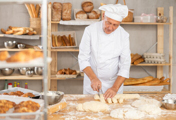 Mature man works in bakery as baker, cuts dough into portions, forms pieces of dough to create buns. Working moment, process of creating croissants in bakery