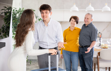 Cheerful young guy greeting wife returning from trip with open arms while friendly elderly parents-in-law standing in background during warm family welcome..