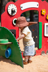 The child is interacting with a colorful play structure that includes a bright red activity panel designed for sand-based play. The scene is set on a sunny day, emphasizing early childhood exploration