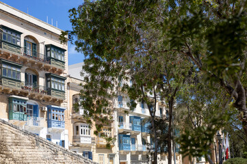 A row of traditional old houses in Valletta, Malta against a dark green foliage of trees lined up in the street.