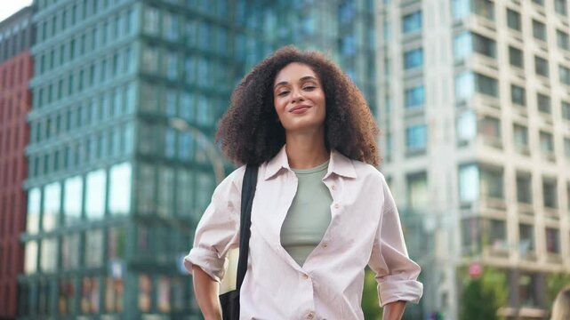 Young pretty woman with curly hair standing confidently outdoors, smiling at modern high-rise buildings. City street. Beautiful happy cheerful female smiling and laughing to camera. Portrait.