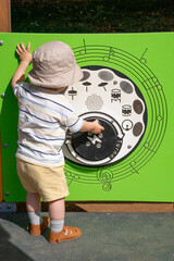 Musical activity panel at an outdoor playground. The child is interacting with bright green sound panel featuring musical notes and drums icons. Scene captures a moment of early sensory exploration. 
