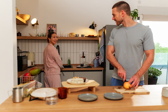 Young man cutting fruit while his partner is washing dishes in their modern and bright kitchen, they're wearing pajamas and enjoying their morning routine together - Powered by Adobe