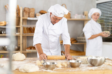 Elderly experienced baker rolls out dough with rolling pin in the kitchen