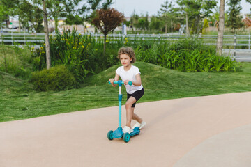 A little curly-haired girl rides a scooter in a city park.