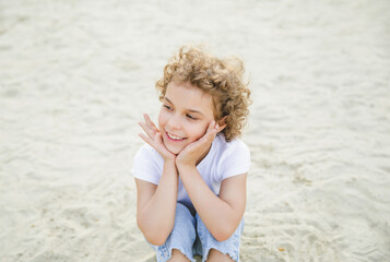 portrait of a little curly cheerful blonde girl sitting on the sand
