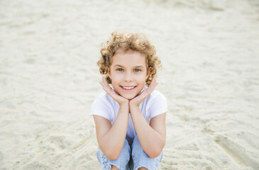 portrait of a little curly cheerful blonde girl sitting on the sand