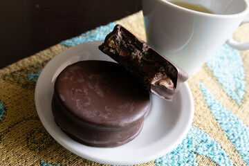 A whole alfajor and a bitten alfajor on a small plate next to a cup of tea