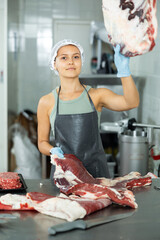 Female employee of local butcher shop divides large huge piece of meat carcass with knife and puts...