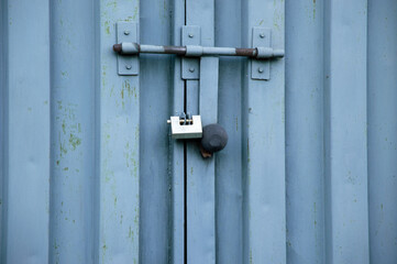 A blue metal gate is secured with a padlock, revealing a narrow gap. The surface shows wear from exposure to the elements