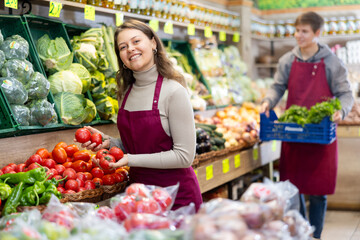 Smiling young saleswoman taking tomatoes from food stall in large grocery store