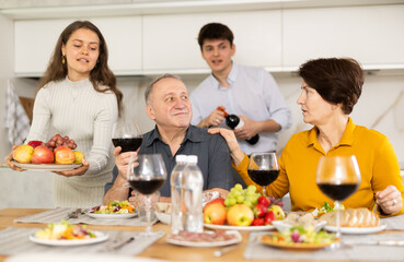 Mother and father visiting young couple, sitting with glass of wine in kitchen. Happy girl serving fruit, excited guy in background