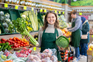 Young woman seller in apron lays out cabbage and celery on counter in vegetable shop