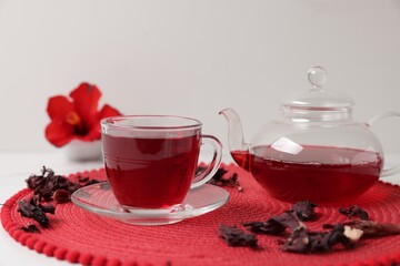 Delicious hibiscus tea in glass cup, teapot and dry roselle sepals on white table, closeup