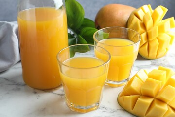 Tasty mango juice in glasses, fresh fruits and green leaves on white marble table, closeup