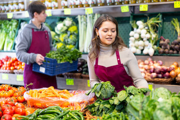 Smiling young saleswoman putting bunch of spinach on counter in large grocery store