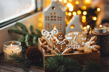 Concept of home christmas or new year celebration, family traditions. Assortment of gingerbread cookies with icing in a vintage wooden box on a window sill with atmospheric lights