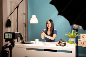 Asian beauty vlogger with lipstick in hand, filming makeup tutorial in studio. Friendly blogger...