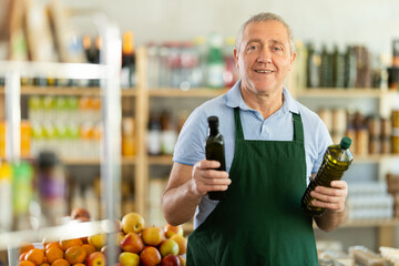 Elderly male seller at counter offering olive oil in bottle in grocery store