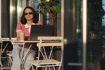 Young woman working on a laptop while enjoying coffee at an outdoor cafe in the midday sun