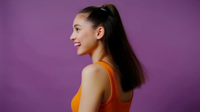  A cheerful young brunette woman with her hair in a ponytail smiles happily while turning her head in profile. Her hair swings against a solid purple background. She wears a bright orange top. 