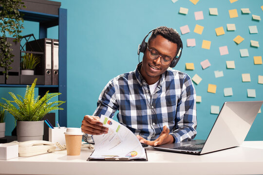 African american financial advisor wearing headset, flipping papers on a clipboard, reviewing company data at desk, planning a business project. Businessman enjoys music during his office hours.