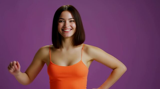  Portrait of a joyful young woman with dark hair smiling and laughing happily. She wears a vibrant orange camisole and gestures thumbs up, isolated on a solid purple background. 