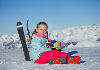 Child in red jacket sits with skis, helmet at side, peaks behind