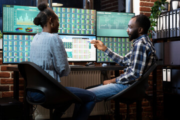 Two forex traders working side by side at desk, tracking stock market trends on multi screen...
