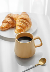 Coffee espresso in yellow cup and fresh croissants on white table near window with morning shadows.
