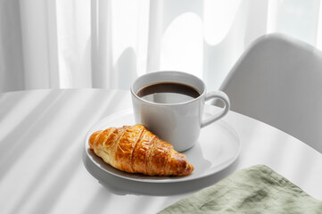 Cup of espresso coffee and fresh croissant on a plate on white table near window