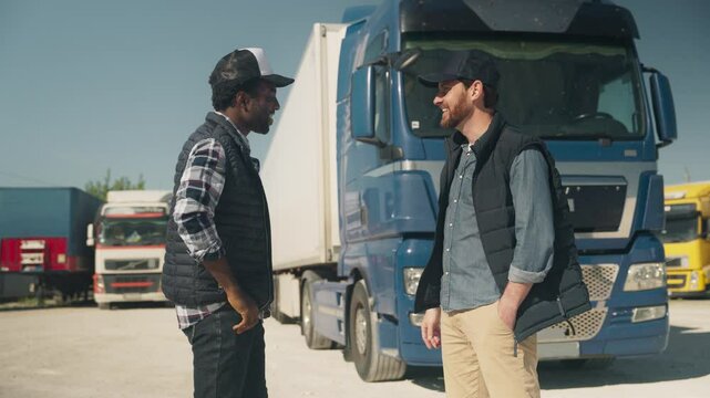 Laughing man in plaid shirt standing near cheerful colleague gesturing with open arms in front of parked truck. Joking while waiting for schedule confirmation. Enjoying light moment before departure.
