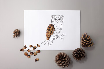 Owl drawing with pine cone scales forming wing on white paper. Studio craft photography on grey background. Autumn forest and DIY education concept.