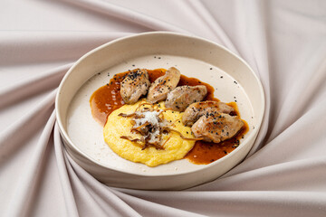 A culinary masterpiece features perfectly cooked chicken pieces drizzled with rich sauce, accompanied by velvety polenta and garnished with delicate herbs, all resting on soft fabric