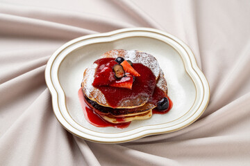 A stack of fluffy pancakes sits invitingly on a decorative plate, adorned with a rich berry sauce and vibrant fresh fruits. The warm hues create a cozy ambiance perfect for brunch