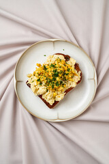 A delicious breakfast features fluffy scrambled eggs garnished with vibrant herbs and spices, elegantly placed atop a slice of toasted bread. The soft fabric background adds a touch of sophistication