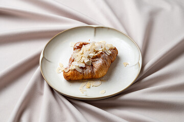 A beautifully baked almond croissant sits elegantly on a fine plate, adorned with nutty flakes atop. Soft, draped fabric provides a warm backdrop, enhancing the culinary charm