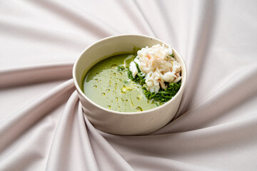 A vibrant bowl of green soup filled with lush herbs, garnished with tender crab meat, rests elegantly on a smooth beige fabric. The inviting colors and textures make it a mouthwatering display