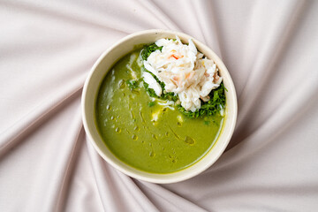 A bowl of vibrant green soup features a silky consistency, topped with tender crab meat and fresh greens, set on a soft fabric backdrop. Ideal for a comforting dining experience