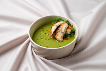 A vibrant green soup is beautifully presented in a smooth round bowl, topped with perfectly grilled chicken. The dish rests on a soft, flowing fabric, enhancing the aesthetic appeal of the meal