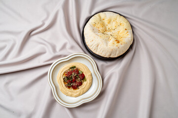 Savor the appealing combination of creamy hummus topped with fresh herbs and spices, paired with fluffy pita bread, all set against a delicate fabric backdrop, inviting culinary exploration