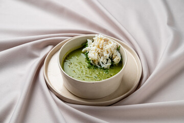 A luxurious bowl of crab soup featuring a vibrant green broth, topped with tender crab meat and accompanied by fluffy white rice. The dish is artfully arranged on a smooth, neutral-toned fabric