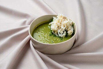 A bowl of vibrant green soup is beautifully garnished with tender crab meat and a sprinkle of fresh herbs, all set against a soft, draped tablecloth