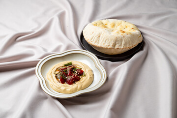 A delightful arrangement featuring creamy hummus garnished with colorful vegetables and herbs, paired with freshly baked pita on a soft, neutral tablecloth. Perfect for sharing