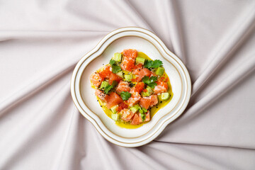 Fresh salmon and creamy avocado are diced and finely mixed, garnished with herbs, placed on a decorative plate against a soft fabric backdrop, exuding a gourmet vibe