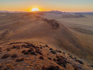 Golden hour in the hills of Carrizo Plain National Monument, California, USA. The sun sets over the rolling landscape, casting a warm glow on the dry grass and shrubs. A serene and peaceful scene.