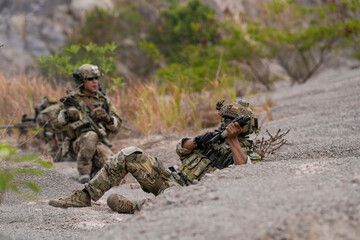 Special forces soldier uses a radio during a tactical operation while red flare smoke spreads across the rocky ground. Team communicates under high-alert battlefield conditions.