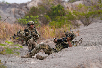Special forces soldier uses a radio during a tactical operation while red flare smoke spreads across the rocky ground. Team communicates under high-alert battlefield conditions.