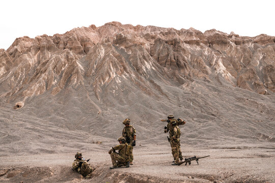 A group of special forces soldiers conduct a briefing in a rugged desert mountain environment. Tactical gear and sniper rifle visible, emphasizing military readiness in harsh terrain.